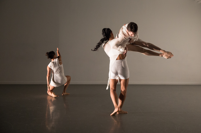 Three women all in white. Two women stand with their back to the audience. One of those women swings another at her armpit. The woman lifted has her legs straight at a ninety degree angle from her hip.